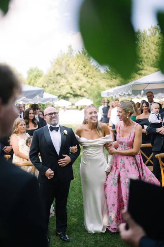 A bride in an off-shoulder white gown walks outdoors with a man in a tuxedo and a woman in a pink floral dress, surrounded by smiling guests at a wedding ceremony. by Justin Salem Meyer