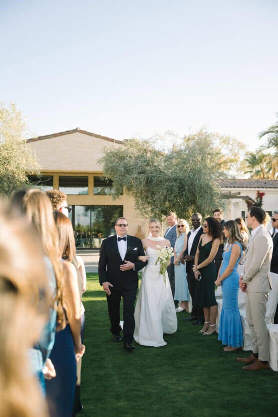 A bride in a white dress holding a bouquet walks down an outdoor aisle on grass, escorted by a man in a tuxedo, while guests stand on either side watching. A building and trees are in the background under a clear sky. by Justin Salem Meyer