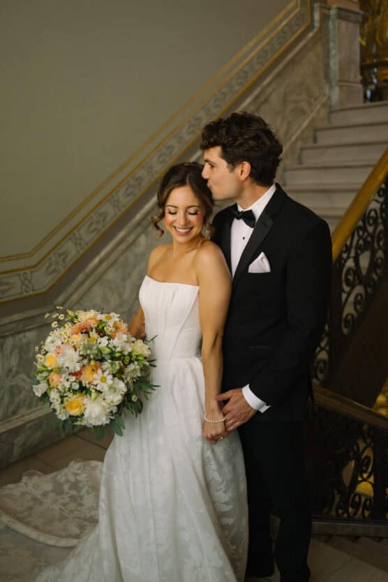 A bride in a white strapless gown holds a bouquet of flowers and smiles while the groom, in a black tuxedo, stands behind her, gently kissing her head. They are on a grand staircase with ornate railings. by Justin Salem Meyer