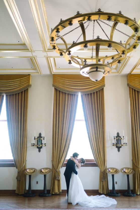A bride and groom embrace and dance in front of tall windows with gold curtains in an elegant room, beneath a large ornate chandelier. by Justin Salem Meyer