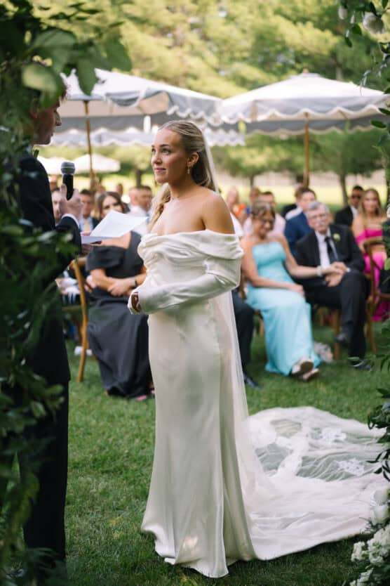 A bride in an off-shoulder white gown stands outdoors facing her partner during a wedding ceremony, with guests seated in the background under umbrellas on a sunny day. by Justin Salem Meyer