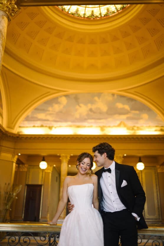 A smiling bride in a white dress stands beside a groom in a black tuxedo under a grand, domed ceiling with warm lighting and ornate architectural details. by Justin Salem Meyer