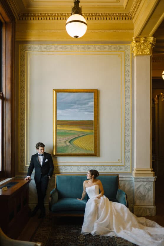 A bride in a white gown sits on a teal sofa while a groom in a black tuxedo leans nearby. They are in an elegant, ornate room with a large landscape painting on the wall behind them. by Justin Salem Meyer