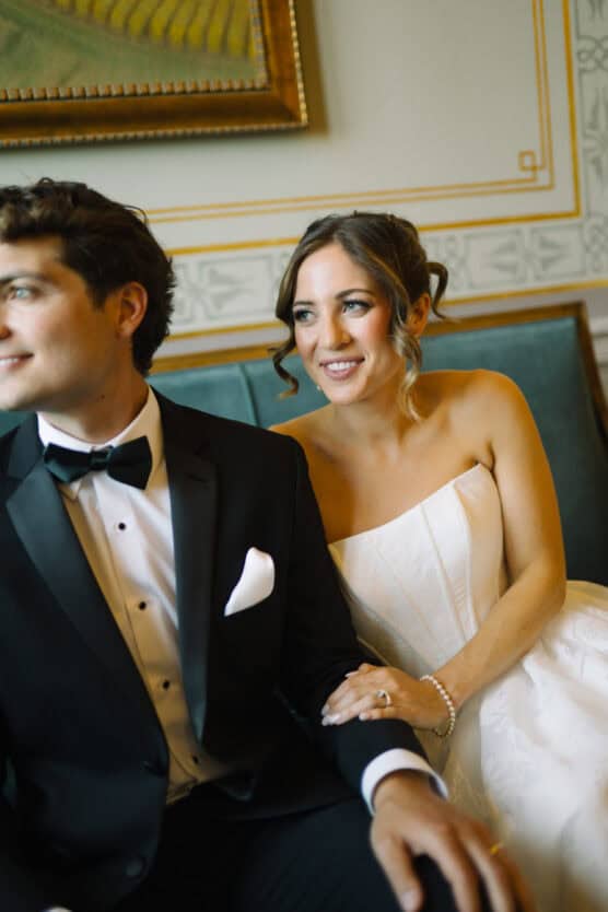A bride in a strapless white gown sits beside a groom in a black tuxedo, both seated on a green bench. The bride smiles while holding the groom’s arm. An ornate wall and framed painting are in the background. by Justin Salem Meyer