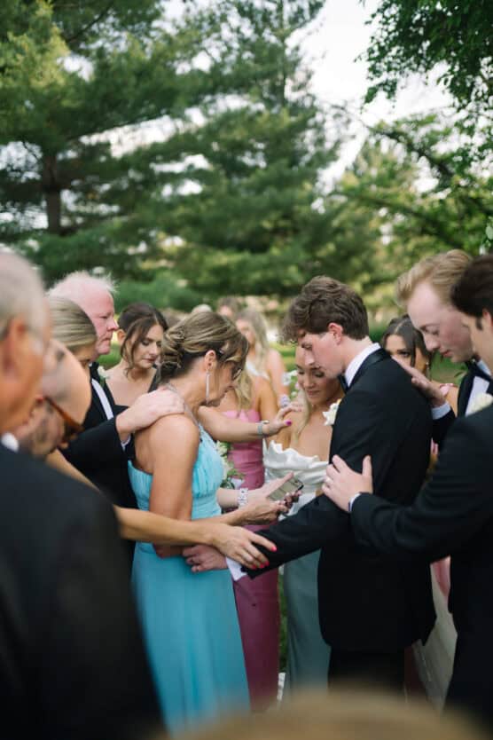 A group of people, dressed formally, gather outdoors around a bride and groom, placing hands on them in a supportive gesture, possibly during a wedding prayer or blessing ceremony. Trees and greenery are in the background. by Justin Salem Meyer