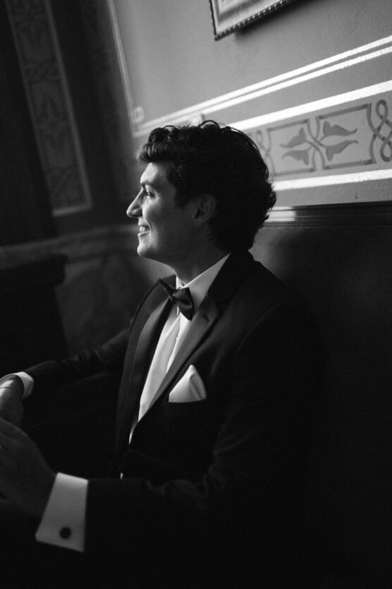 A man in a tuxedo with a bow tie sits and smiles, looking to the side in a dimly lit, ornately decorated room. The image is in black and white. by Justin Salem Meyer