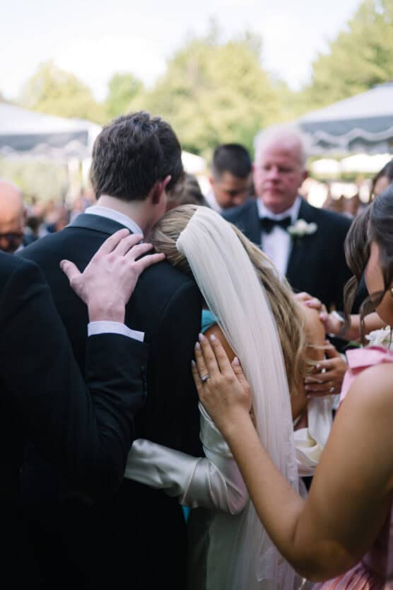 A bride in a white dress and veil leans on a groom in a suit while surrounded by loved ones offering comfort and support at an outdoor wedding ceremony. by Justin Salem Meyer