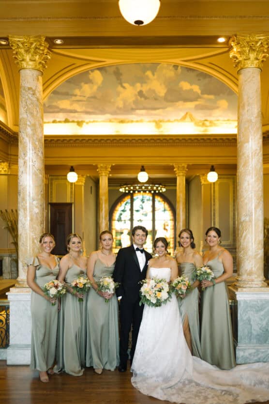 A bride and groom stand together, smiling, flanked by six bridesmaids in light green dresses, holding bouquets. They are indoors, in an elegant hall with marble columns and a large, decorative window behind them. by Justin Salem Meyer