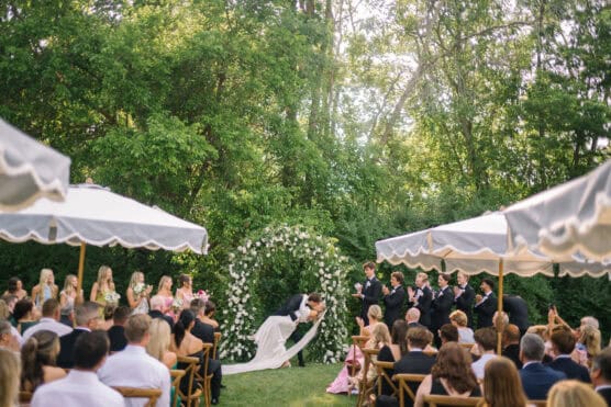 A bride and groom kiss under a flower arch during an outdoor wedding ceremony surrounded by guests seated under umbrellas, bridesmaids in pink dresses, and groomsmen in black suits. Lush green trees fill the background. by Justin Salem Meyer