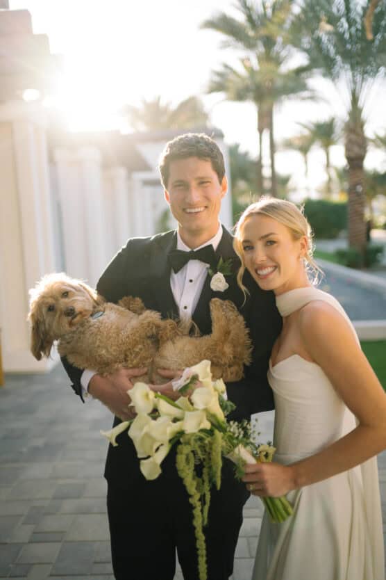 A smiling bride and groom pose outdoors in wedding attire, with the groom holding a fluffy brown dog in his arms. The bride holds a bouquet of white flowers. Palm trees and sunlight are in the background. by Justin Salem Meyer