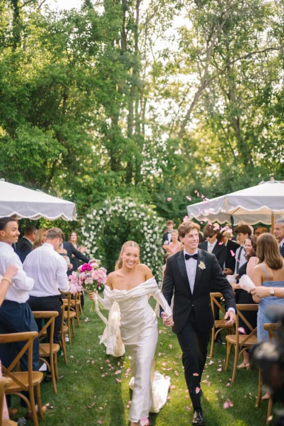 A joyful bride and groom walk hand in hand down an outdoor aisle, smiling as guests throw flower petals. The bride holds a bouquet and wears an off-shoulder white gown; the groom wears a tuxedo. Trees and tents are in the background. by Justin Salem Meyer