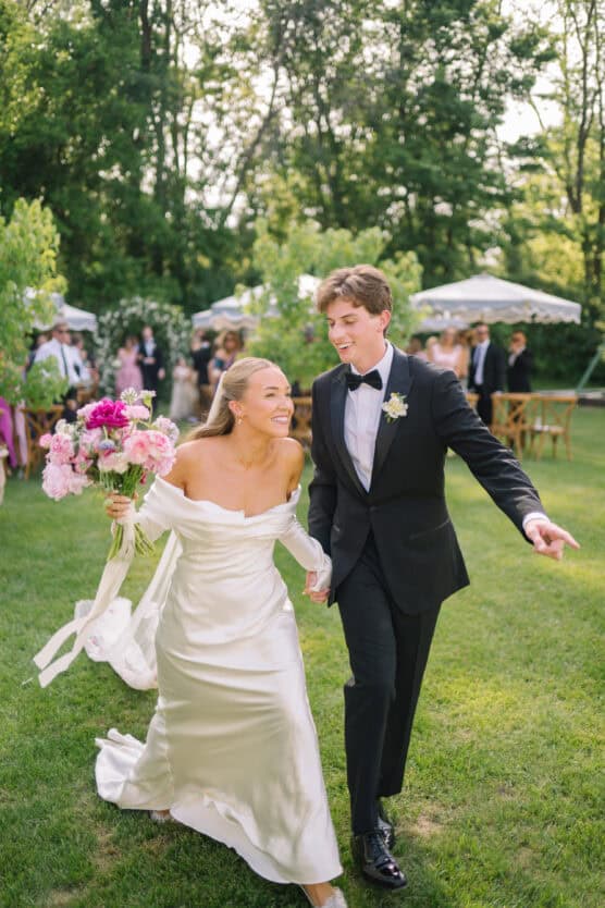 A joyful bride in an off-the-shoulder white dress holds a bouquet of pink flowers and walks hand-in-hand with a smiling groom in a black tuxedo outdoors, surrounded by greenery and wedding guests. by Justin Salem Meyer