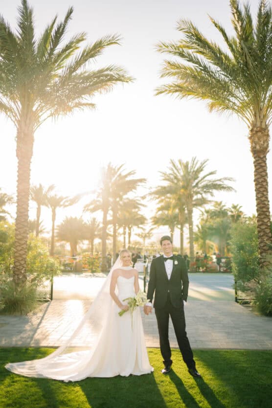 A bride in a white gown and veil stands holding hands with a groom in a black tuxedo. They are outdoors, surrounded by tall palm trees, with sunlight streaming behind them. by Justin Salem Meyer