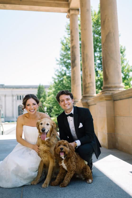 A bride and groom in wedding attire kneel outdoors on stone pavement, smiling beside two happy dogs. Tall columns and green trees are visible in the background on a sunny day. by Justin Salem Meyer