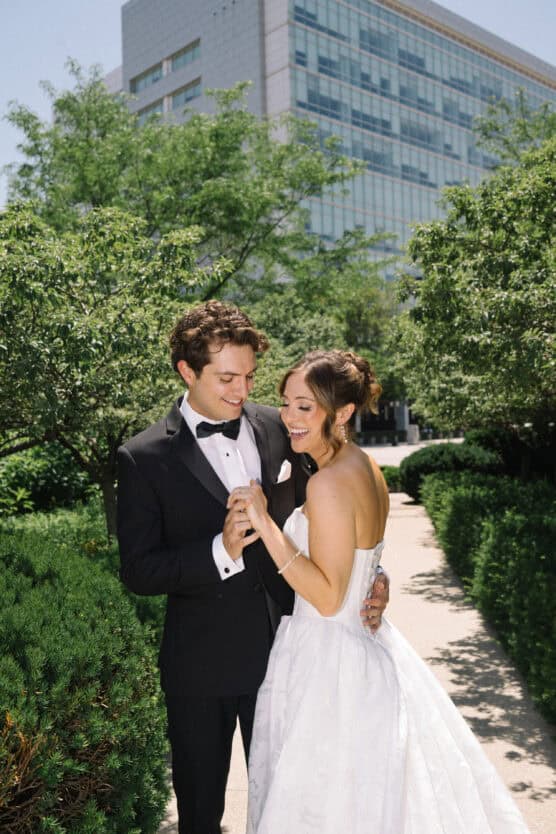 A bride in a strapless white gown and a groom in a black tuxedo smile and hold hands while standing on a garden path, with greenery and a modern building in the background. by Justin Salem Meyer