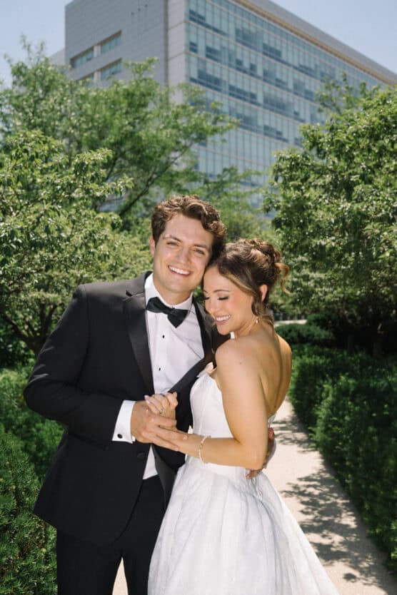 A smiling couple dressed in formal wedding attire stands close together on a sunlit outdoor path, surrounded by greenery, with a modern building in the background. by Justin Salem Meyer