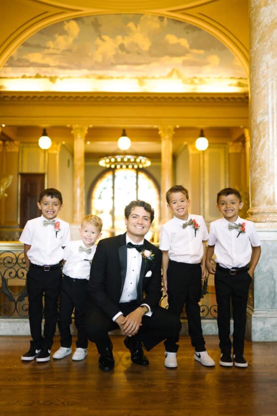 A man in a tuxedo crouches, smiling, surrounded by four young boys in white shirts, dark pants, and bow ties. They stand in an ornate hall with marble columns and warm lighting. by Justin Salem Meyer