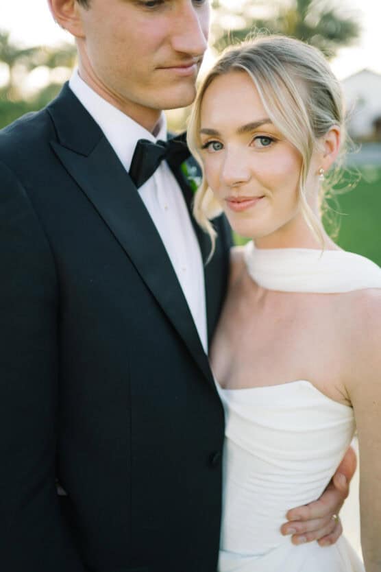 A bride in a white dress and a groom in a black suit stand closely together outdoors. The bride smiles softly at the camera, while the groom looks down, partially out of frame. by Justin Salem Meyer