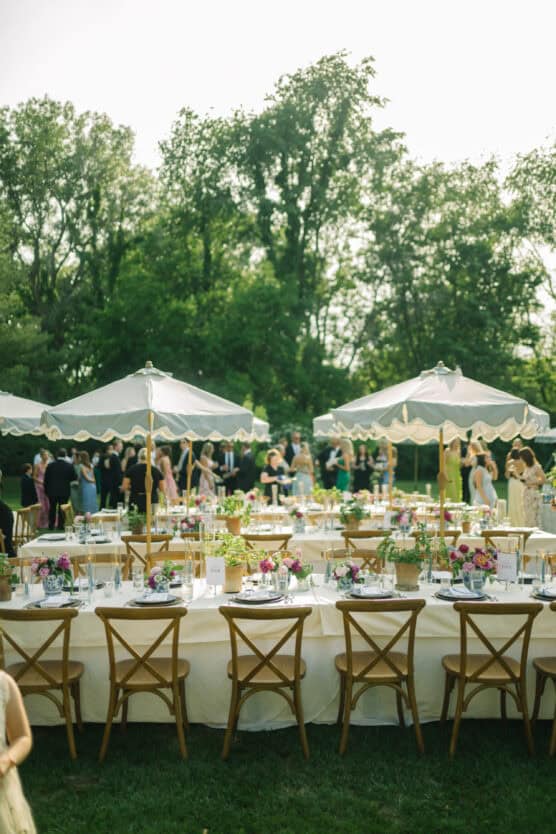 Outdoor garden party with tables set for dining, wooden chairs, white tablecloths, floral centerpieces, and large white umbrellas. People are mingling in the background among lush green trees. by Justin Salem Meyer