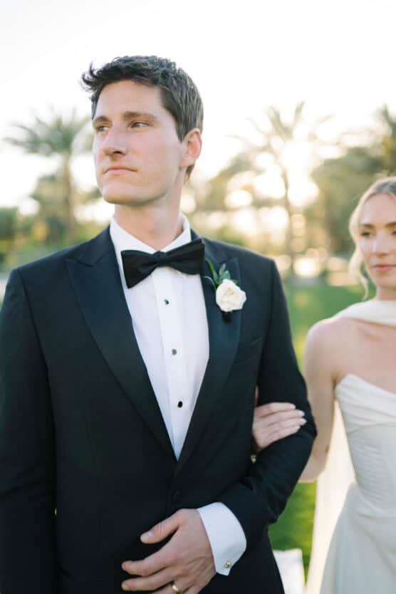 A groom in a black tuxedo with a white rose boutonniere stands outdoors with a bride in a white dress holding his arm, palm trees and sunlight in the background. by Justin Salem Meyer