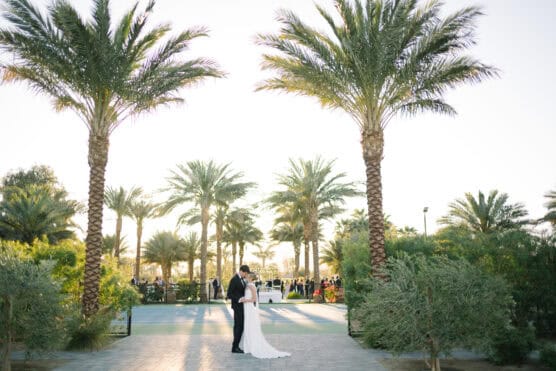 A bride and groom stand closely together on a pathway surrounded by lush palm trees, with sunlight streaming through the leaves and guests visible in the background. by Justin Salem Meyer