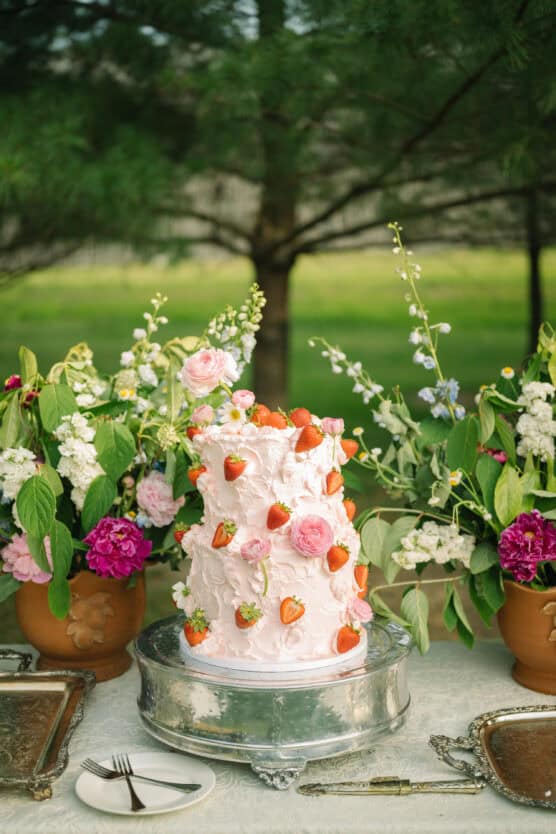 A pink, tiered cake decorated with strawberries and roses sits on a silver cake stand between two large floral arrangements on an outdoor table. A cake knife and server are placed nearby. by Justin Salem Meyer