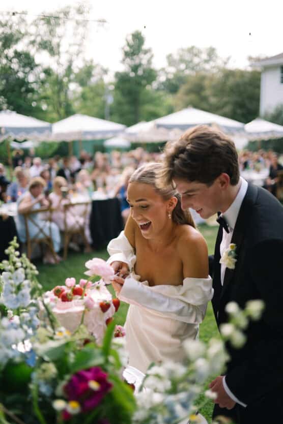 A bride and groom laugh joyfully while cutting a wedding cake decorated with strawberries and pink flowers at an outdoor reception, with guests seated in the background. by Justin Salem Meyer