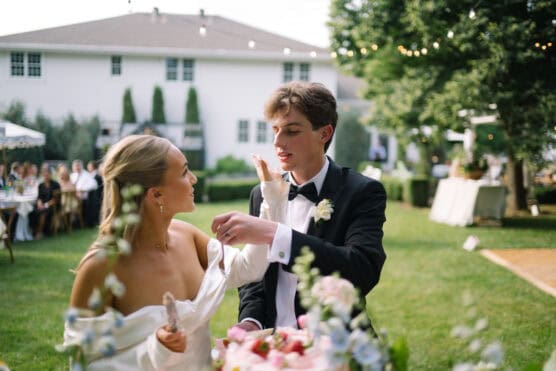 A bride and groom in formal attire feed each other cake outdoors during their wedding reception, surrounded by greenery and decorated tables. by Justin Salem Meyer