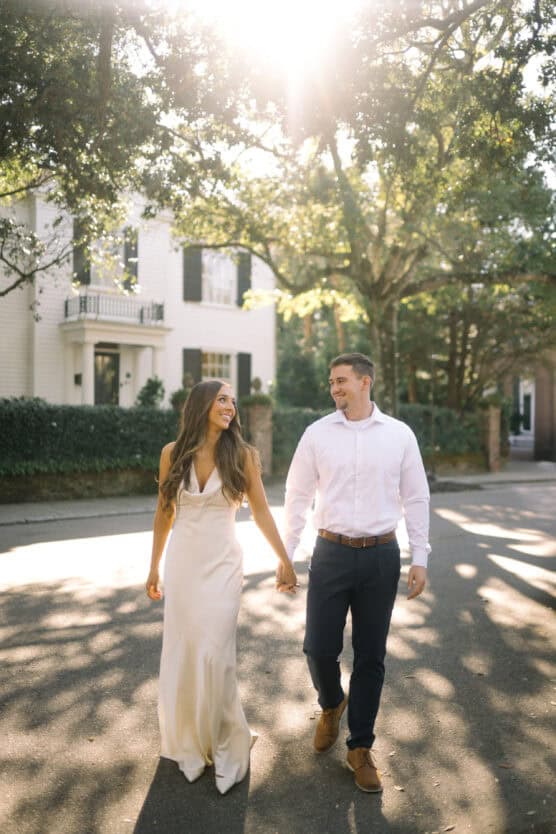 A couple walks hand in hand down a sunlit street, smiling at each other. The woman wears a long white dress, and the man wears a white shirt and dark pants. Trees and a white house are in the background. by Justin Salem Meyer