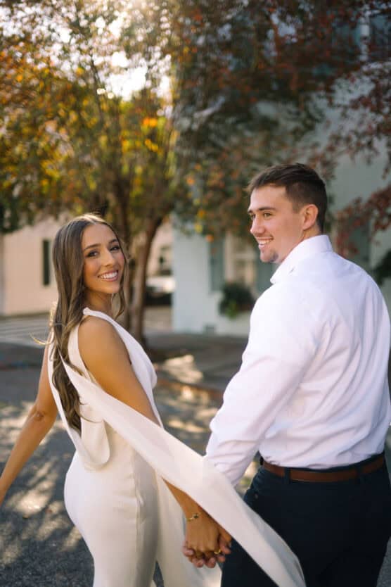A smiling couple holds hands and looks back at the camera while walking outdoors. The woman wears a white dress, and the man wears a white shirt. Sunlight filters through trees in the background. by Justin Salem Meyer