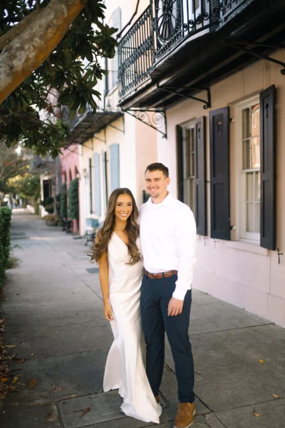 A smiling couple stands on a sidewalk beside pastel-colored buildings; the woman wears a white dress and the man wears a white shirt with dark pants. Trees and iron balconies are visible in the background. by Justin Salem Meyer
