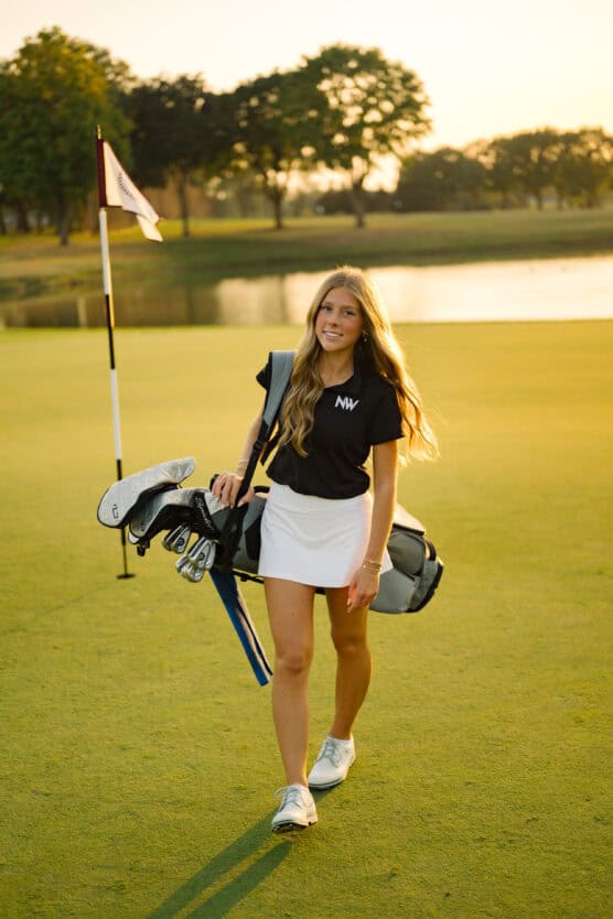 A young woman with long blonde hair walks on a golf course carrying a golf bag, wearing a black shirt and white skirt, with a flag and pond in the background at sunset. by Justin Salem Meyer