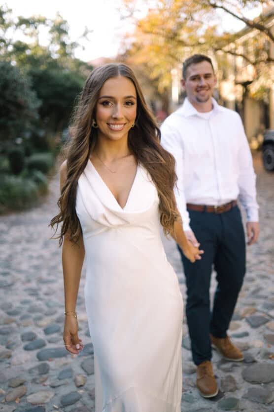 A woman in a white dress smiles and walks ahead, holding hands with a man in a white shirt and dark pants on a cobblestone street with greenery and trees in the background. by Justin Salem Meyer