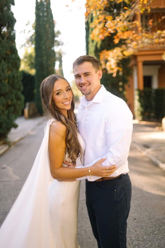 A smiling couple stands close together outdoors, dressed in semi-formal attire; the woman wears a white dress and the man wears a white shirt and dark pants, with trees and a sunlit building in the background. by Justin Salem Meyer