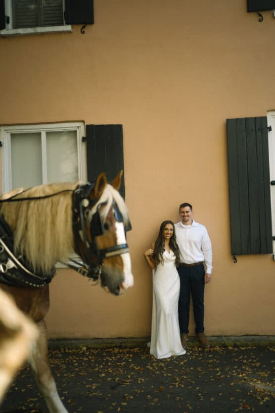 A couple in formal attire stands against a peach-colored wall with black shutters, smiling at the camera. In the foreground, a blond horse in harness partially blocks the view. Fallen leaves cover the ground. by Justin Salem Meyer