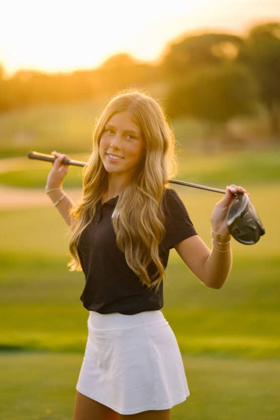 A young woman with long, wavy hair stands on a golf course at sunset, holding a golf club across her shoulders. She wears a black shirt and a white skirt, smiling softly at the camera. The background is softly blurred. by Justin Salem Meyer