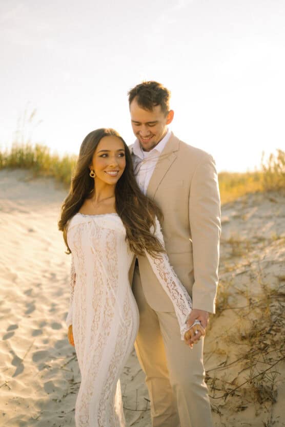 A couple stands on a sandy beach at sunset, holding hands. The woman wears a long, white lace dress and has long brown hair, while the man wears a light beige suit. Both are smiling and looking content. by Justin Salem Meyer
