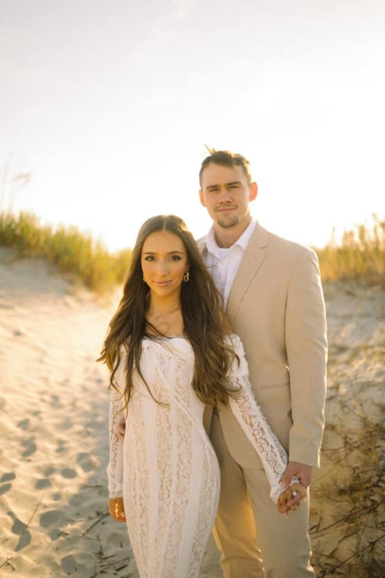 A couple stands on a sandy beach at sunset, holding hands. The woman wears a long white lace dress; the man is dressed in a light beige suit. Grasses and sunlight create a warm, natural background. by Justin Salem Meyer