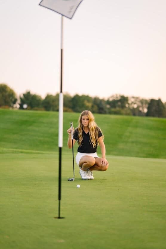 A young woman in a black shirt and white skirt kneels on a golf green, concentrating as she lines up a putt near the hole, with a golf flag and lush green grass in the background. by Justin Salem Meyer
