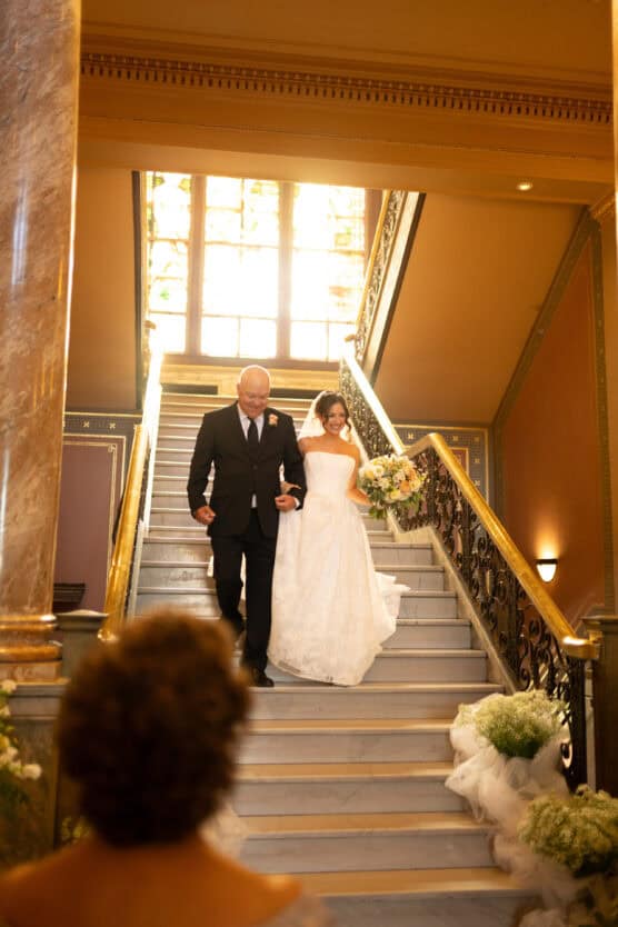 A bride in a white gown and a man in a suit walk arm-in-arm down a marble staircase, with sunlight streaming through a window behind them. A person stands at the bottom of the stairs, facing them. by Justin Salem Meyer