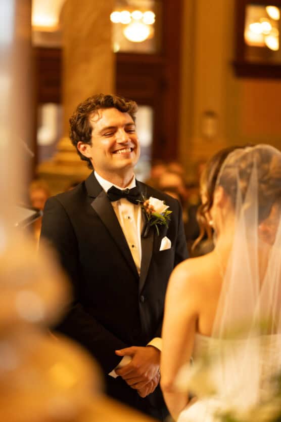 A groom in a tuxedo smiles warmly at his bride, who is wearing a veil, during a wedding ceremony in an elegant, warmly lit venue. by Justin Salem Meyer