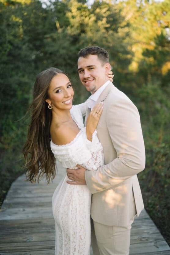 A woman in a white lace dress and a man in a beige suit embrace and smile at the camera while standing on a wooden path outdoors, surrounded by greenery. by Justin Salem Meyer