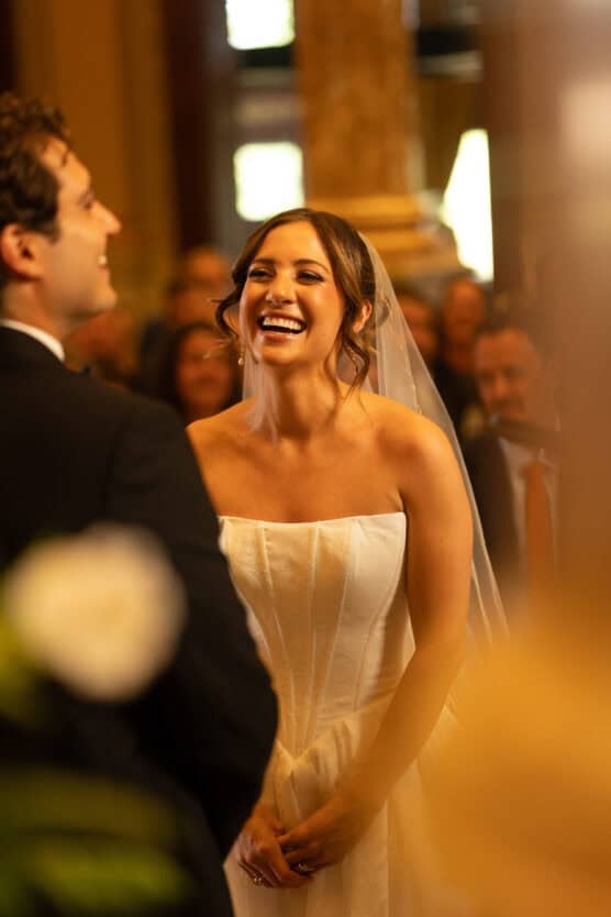 A bride wearing a white strapless gown and veil smiles joyfully during a wedding ceremony, standing across from the groom in a suit, with guests seated in the softly lit background. by Justin Salem Meyer