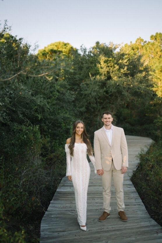 A couple stands on a wooden boardwalk surrounded by lush green trees. The woman wears a long white lace dress and heels, and the man is in a beige suit with a white shirt and brown shoes. They are holding hands and smiling. by Justin Salem Meyer