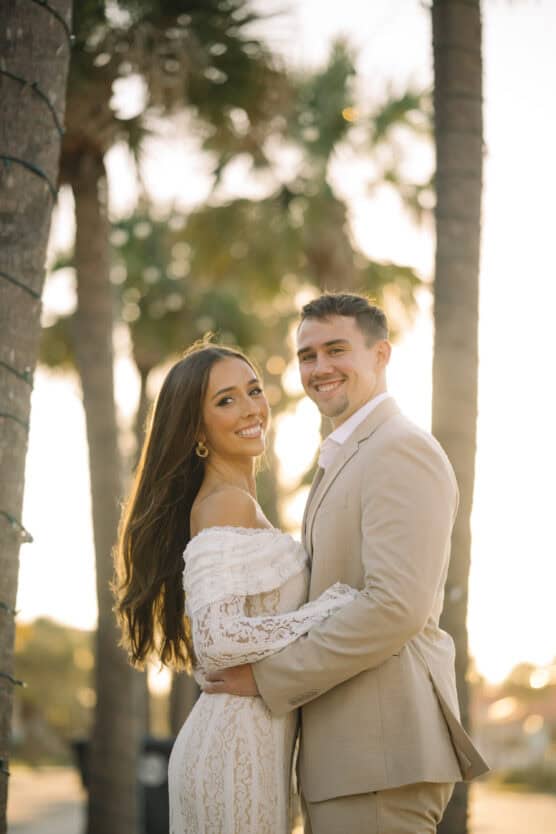 A couple stands outdoors among palm trees at sunset. The woman wears a white lace off-shoulder dress and the man wears a beige suit. They are smiling and embracing each other, with warm sunlight in the background. by Justin Salem Meyer