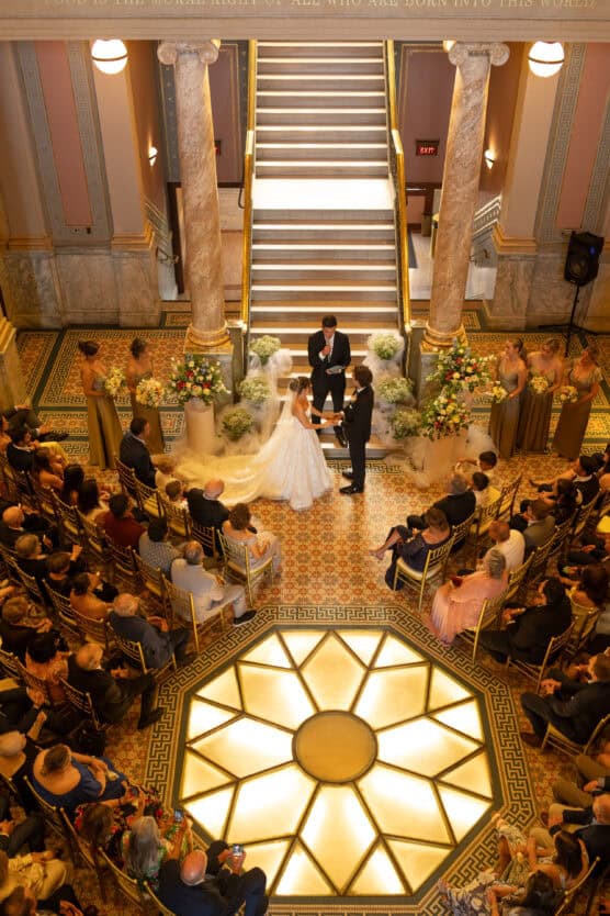 A bride and groom stand with an officiant at the altar in a grand hall, surrounded by guests and a wedding party on either side. The ceremony takes place beneath a large staircase and ornate columns. by Justin Salem Meyer