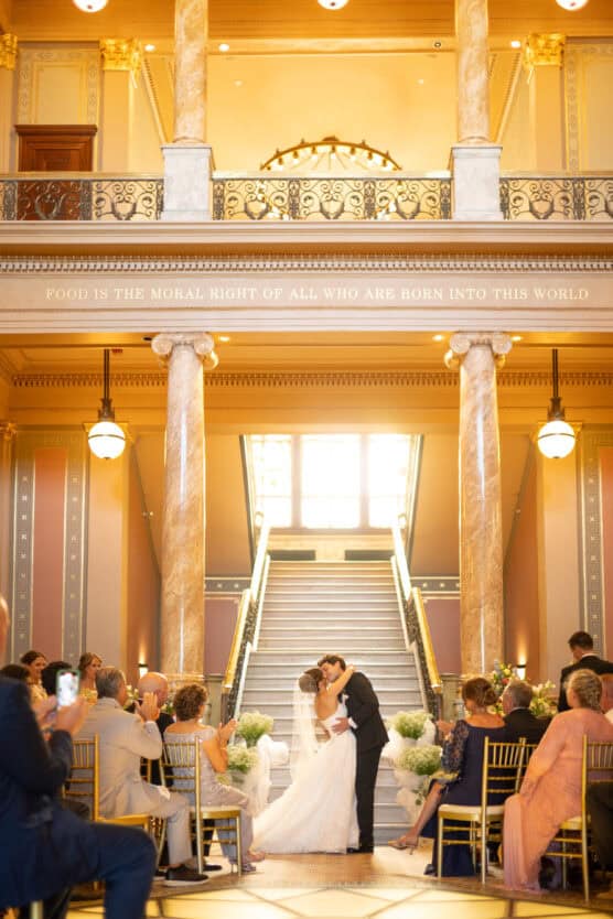 A bride and groom share a kiss at the end of the aisle in a grand hall with marble columns, surrounded by seated guests, under a balcony inscribed with “FOOD IS THE MORAL RIGHT OF ALL WHO ARE BORN INTO THIS WORLD.”. by Justin Salem Meyer