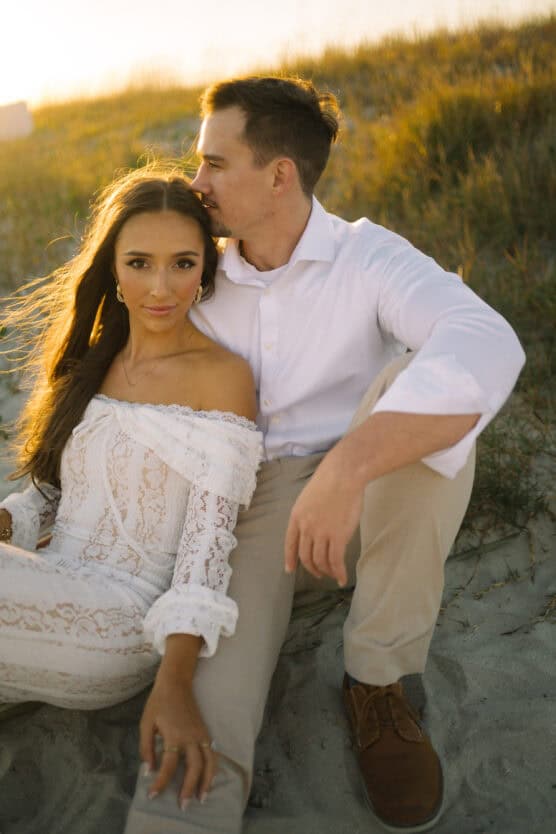 A couple sits on the sand at sunset. The woman wears a white off-shoulder lace dress, and the man in a white shirt and beige pants sits beside her, embracing her affectionately. Grassy dunes are in the background. by Justin Salem Meyer