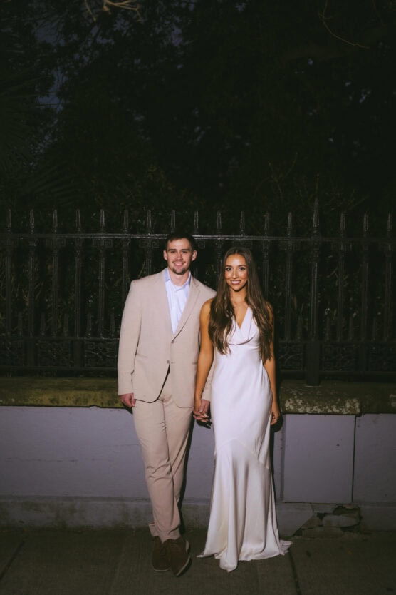 A couple dressed formally, with the man in a beige suit and the woman in a white gown, stand holding hands and smiling in front of a black iron fence at night. by Justin Salem Meyer