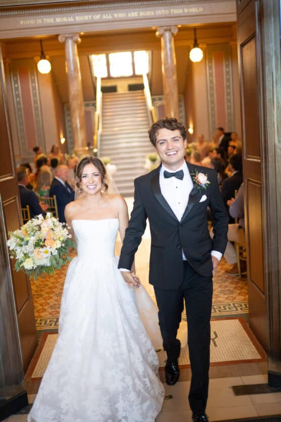 A bride in a white strapless gown and a groom in a black tuxedo walk hand in hand down an aisle, smiling, with wedding guests seated on either side in an elegant hall with columns and stairs in the background. by Justin Salem Meyer